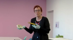 A white person with red hair is wearing protective gloves to hold an ornate gold blood transfusion set, which they are displaying towards the camera, with the workshops online participants able to see