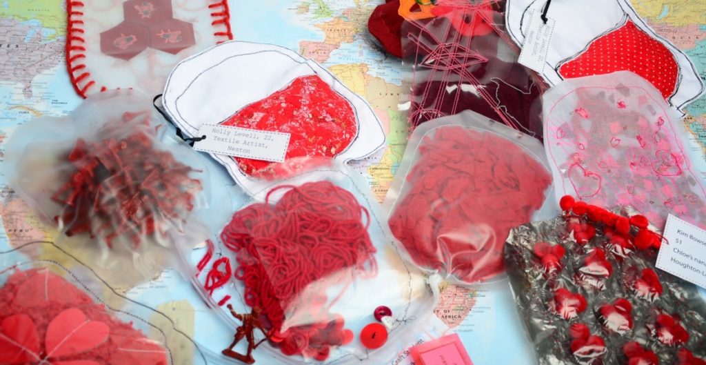 An image of some of the blood bags created as part of the Blood Bag Project. They are made of a diverse selection of materials, many of them are clear plastic blood bags with materials such as red string or red pieces of paper inside them to resemble blood, others are more abstract, some contain images of red hearts on. They are sat on an world map, to indicate the many places across the world that blood bags have been made for the project.