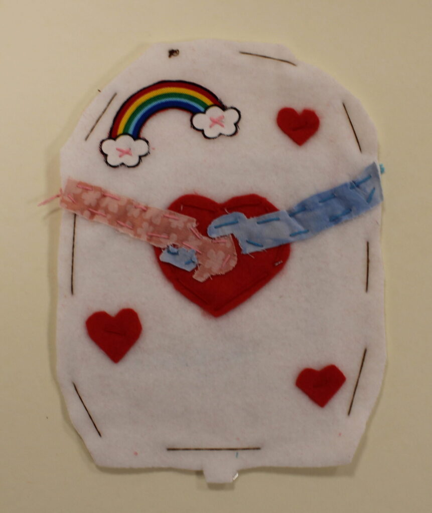 A white felt blood bag featuring two people shaking hands (only their arms and hands are visible). Their hands are being shaken over a red love heart. There are three other red love hearts on the image and a fabric rainbow.